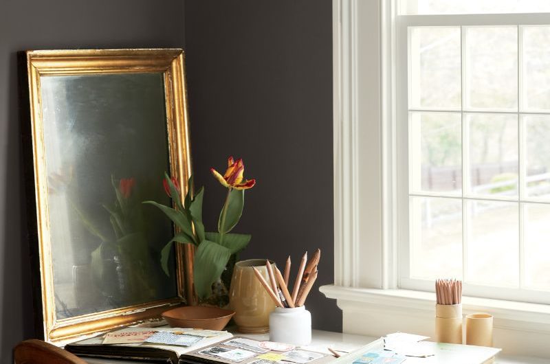 A study with dark brown wall paint, a white desk, vintage chair, and antique mirror.