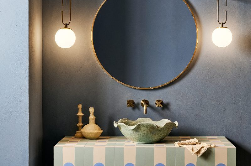 A blue bathroom with geometric tiles, a quirky basin, large circular mirror and wall lights.