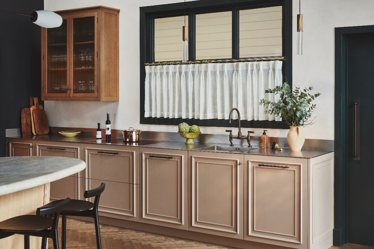 A cooking and sink run in a custom kitchen, with decorative cabinet doors, stainless steel worktops, brass tap, and a cafe curtain on the window above the wash zone.