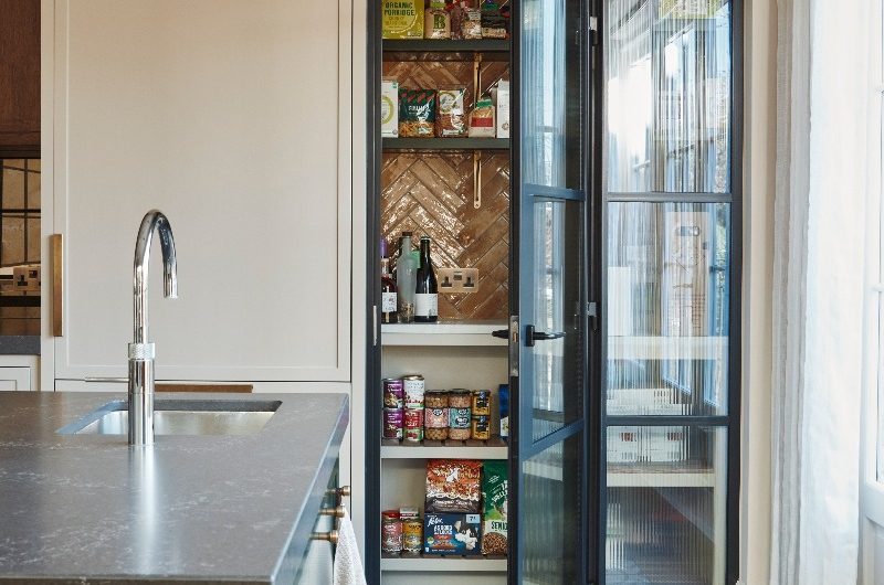A walk-in pantry with glossy tiles, open shelves, steel-framed fluted glass doors, next to sleek neutral cabinetry.