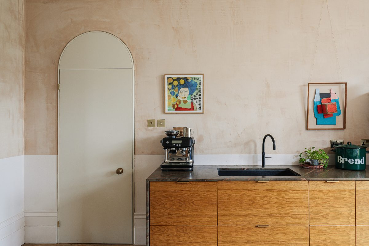 A sink zone with wood units, dark work surfaces, a raw plaster finish on the walls, and a curved door leading to an adjoining room.