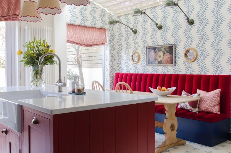 A contemporary kitchen design with a timeless feel, with white and blue wallpaper, a dark red island and an adjoining dining nook with a vibrant red upholstered banquette and wood table.