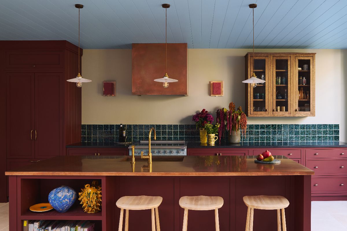 A characterful scheme with burgundy cabinetry, copper and black stone work surfaces, a wood wall unit, green tiles, gold tap, copper extractor, and a blue painted ceiling matching the range cooker.