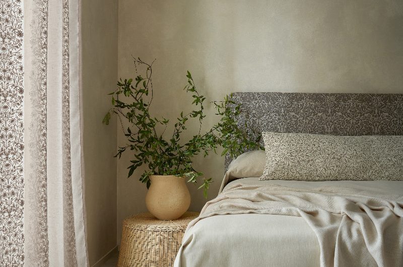 A calm bedroom design with limewash paint, a wood bed frame with an upholstered headboard, neutral linen, rattan side table with a pot and greenery on top of it.