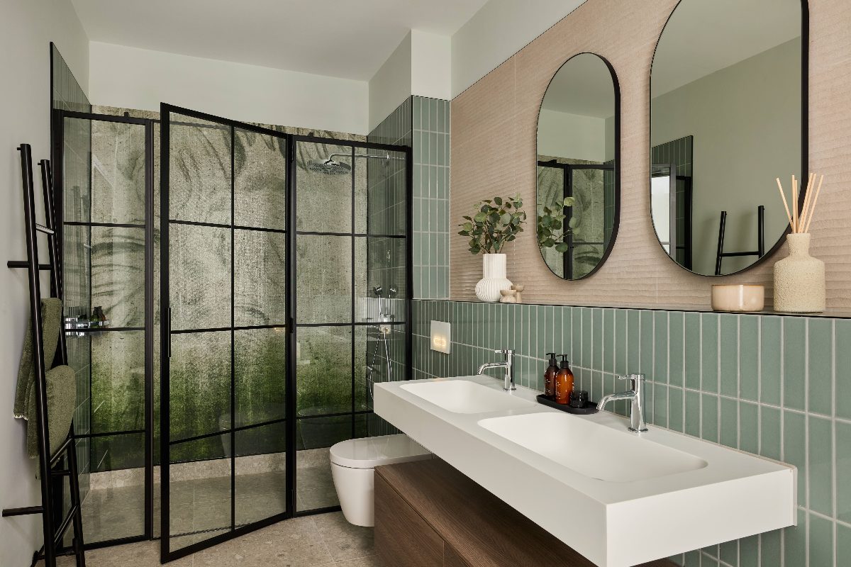 A pink and green couple's bathroom with a double wall-mounted basin, tactile tiles, and a shower zone with a black-frame enclosure and nature-inspired mural.