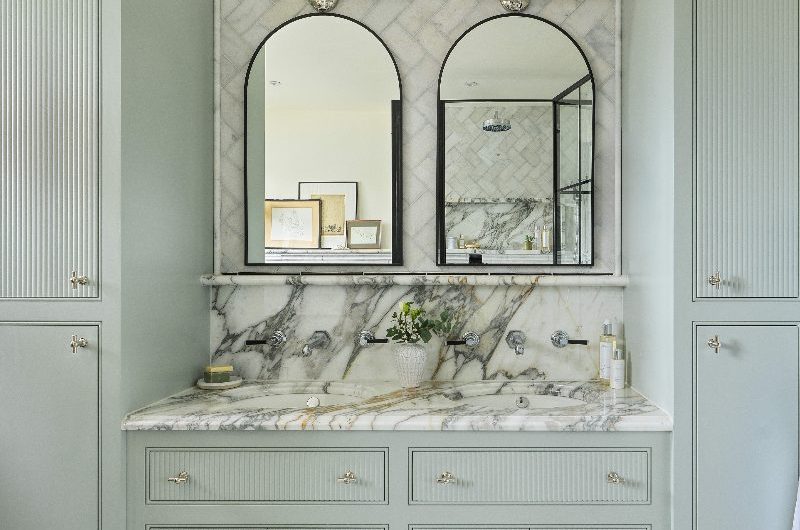 A pale blue bathroom with a double vanity unit with inset basins, marble surfaces, and matching mirrors, lighting and brassware, with wall reeded cabinets either side.