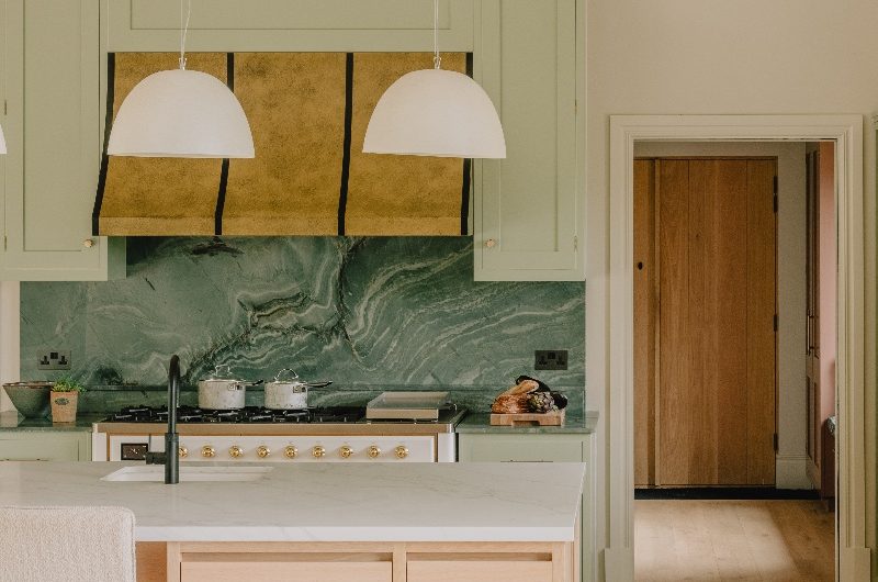 A modern kitchen design with a central island and a run of cabinets behind with a statement range, metallic extractor canopy and green marble work surfaces.