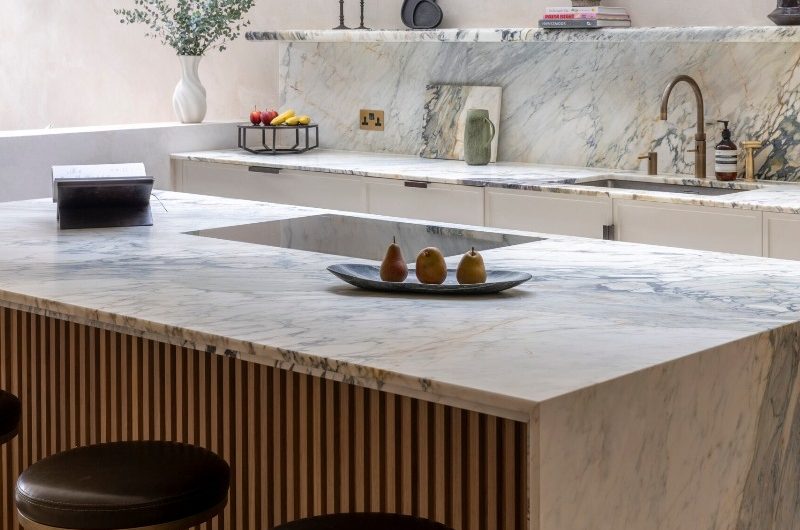 A contemporary cooking space with a Breccia Capraia waterfall unit and matching worktop, splashback, and floating shelf, with plaster on the wall and a long skylight.