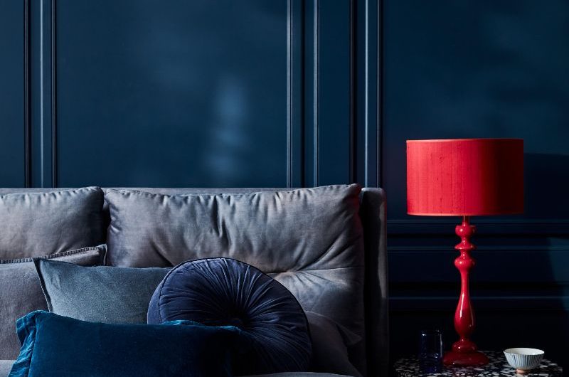 A colour drenched bedroom in blue with timeless panelling, tartan throw, and terrazzo side table with a red table lamp.
