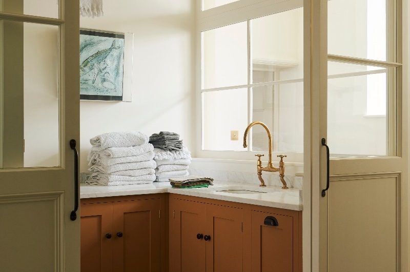 A bright utility room with orange base cabinets, worksurfaces, glass doors, with terracotta flooring, and pendant lighting.