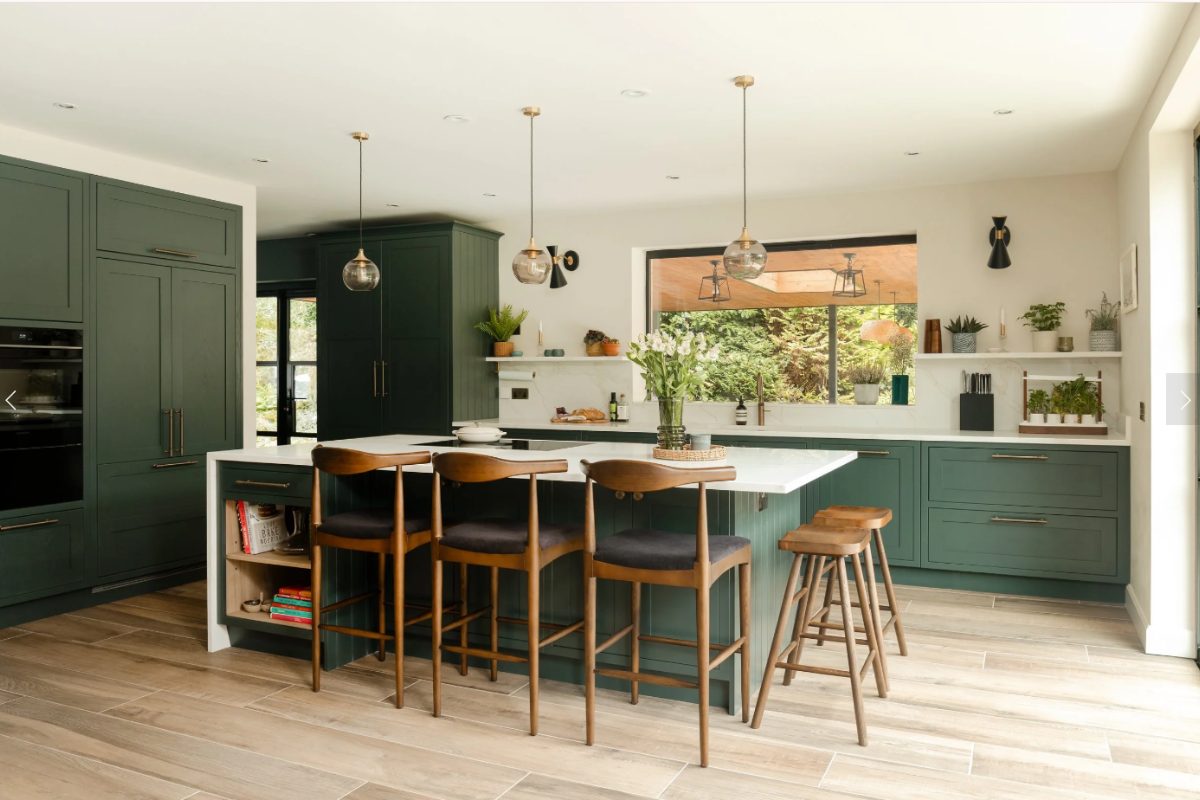 A green kitchen design with classic cabinetry with brass hardware, a big window above the sink area, and a central island with storage, seating, worktop space and a built-in hob.