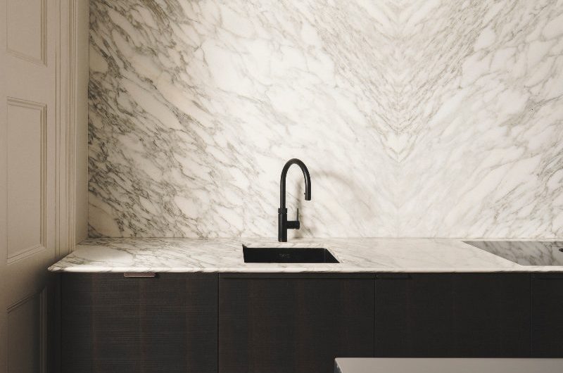 Closeup at a sink zone with grainy wood base cupboards, bookmatched marble spashbacks and matching worktops, a black sink and tap, and elegant cornicing at the top.
