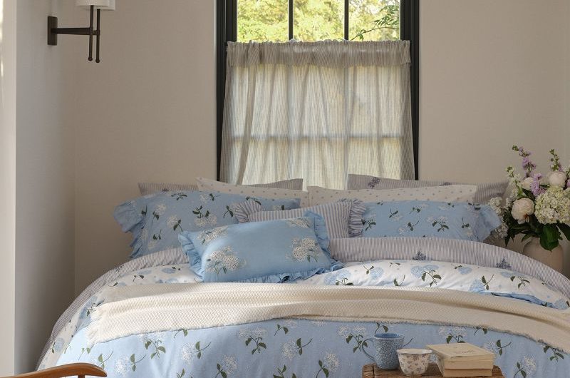 A bright sleep space with airy curtains, blue floral spring bedding and a wood stool.
