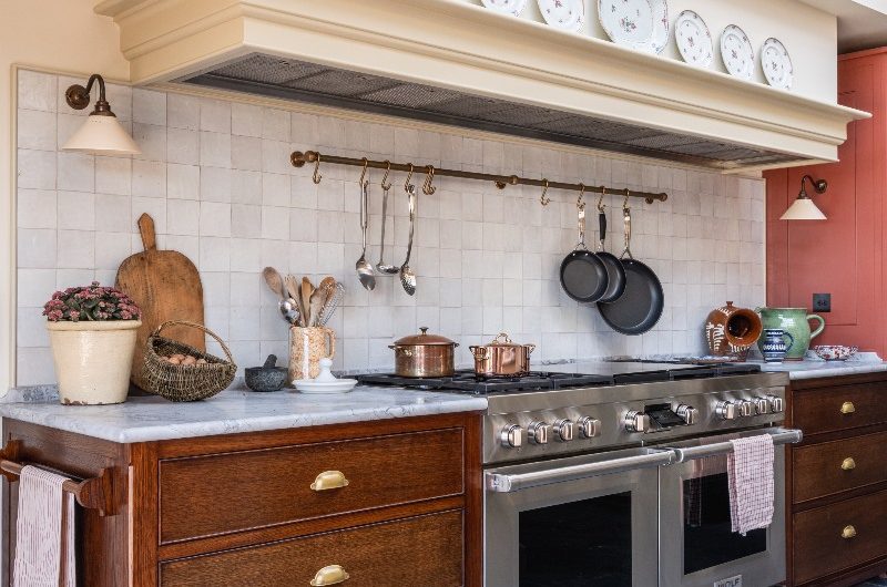 A cooking run in a classic kitchen with wood cabinets on either side of the range cooker, an extractor concealed with a canopy, marble wotkops, and zellige wall tiles.