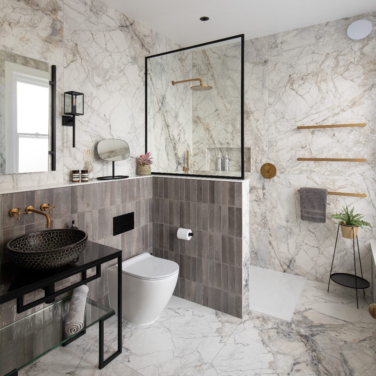 Shower design with a half wall, dark neutral stacked metro tiles, marble wall and floor tiles, and gold and black fittings and fixtures.