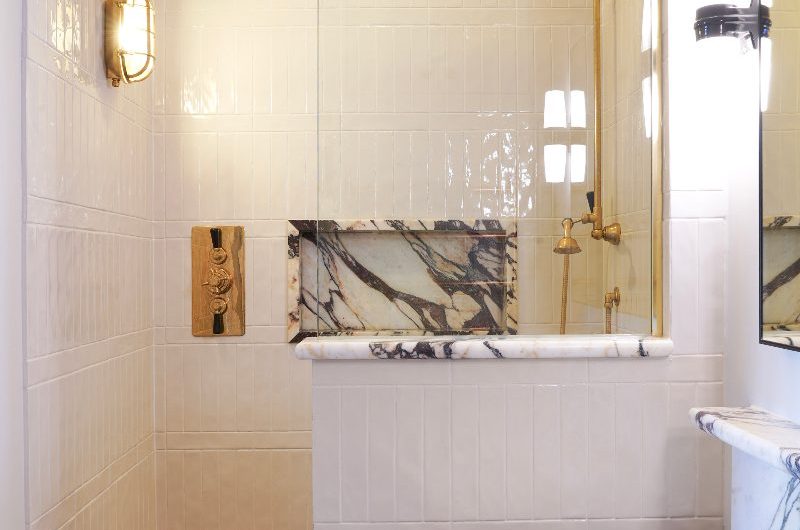 A small boutique bathroom with a half wall with a glass panel, pale pink and glossy wall tile, marble niche and vanity unit, and gold brassware.