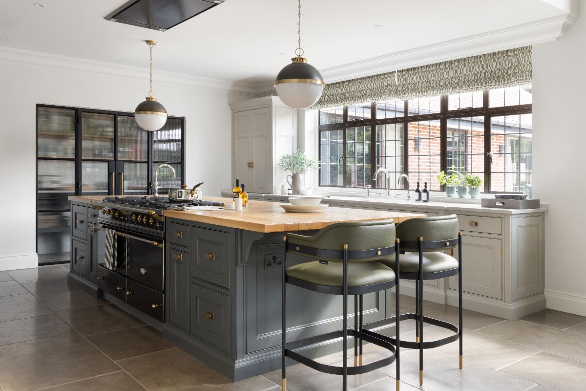 An Arts and Crafts kitchen design with a mix or light and dark grey cabinets, stone flooring, wood worktop on the working island, and lighter surfaces on the sink zone.