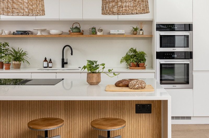 A Scandi scheme with tall, white handleless cabinetry, a central island, a wood open shelf with decorative accessories, and rattan pendant lights.