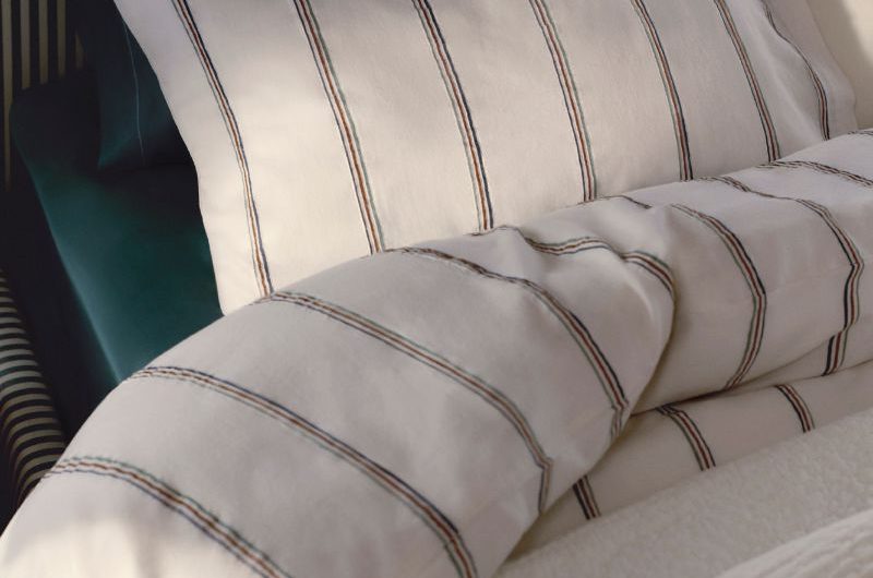 Closeup at a white, striped linen set with dark teal fabrics underneath and a striped headboard.