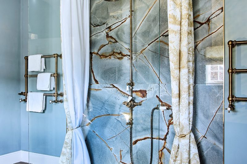 A luxurious space with large marble tiles, blue wall paint, bronze radiators, and an open shower with a sleek panel and patterned shower curtains.