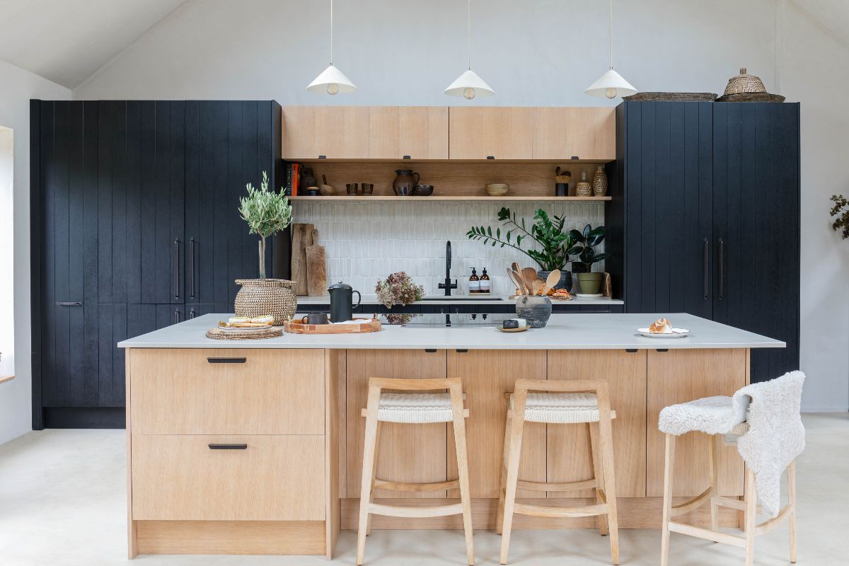 View of a Japandi kitchen with dark timber cupboard hosing a fridge-freezer and pantry on either side of the sink zone, with a central island with storage, seating and a hob.