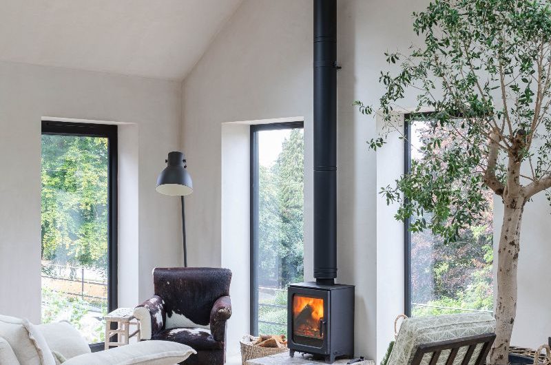 A sitting area with a log burner, cosy seating, tall windows letting light in, and microcement flooring.