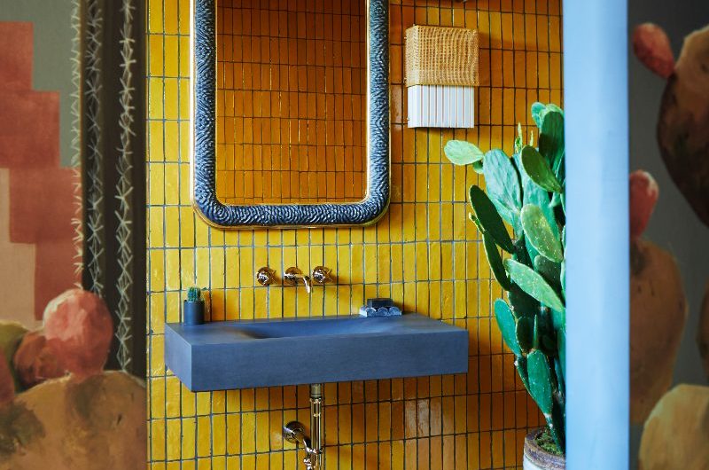 An exotic bathroom layered in colour and texture with yellow wall and floor tiles, blue concrete basin, quirky mirror, and patterned wallpaper.