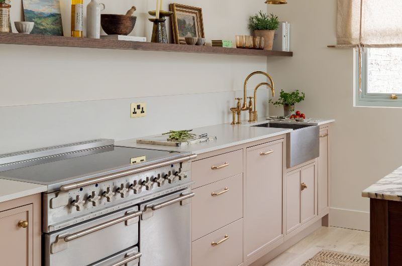 A light pink run of cabinets, with mixed finishes, such as a stainless steel range cooker, brass taps and lighting, and polished brass handles, with dark wood open shelving, and light wood flooring.