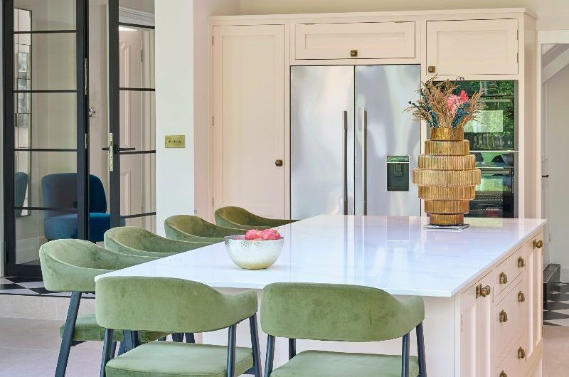 A pink kitchen island with green bar stools and modern, black-framed, indoor partitions which lead to other rooms in the house.
