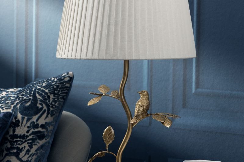 Closeup at a table lamp with a brass base and white shade, with blue timeless panelling in the background, perfect for the Wuthering Heights aesthetic.