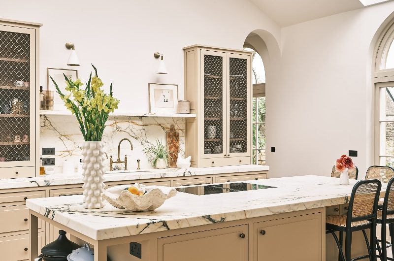 A timeless, neutral space with marble surfaces, a central island with open and closed storage, seating, and a hob, with a sink area behind it and mesh-fronted cabinet doors.