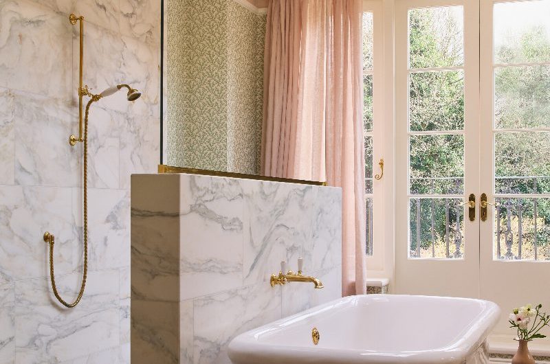 A luxurious bathroom design with a semi-open shower with marble tiles, gold brassware, and a half wall with a freestanding tub positioned against it.