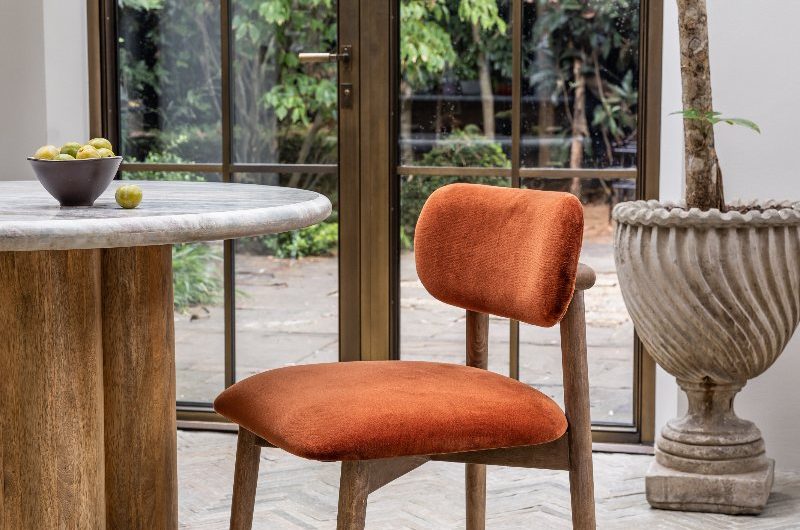 A dining room with a large curved window, terracotta flooring, a modern table and a chair with orange velvet seating.
