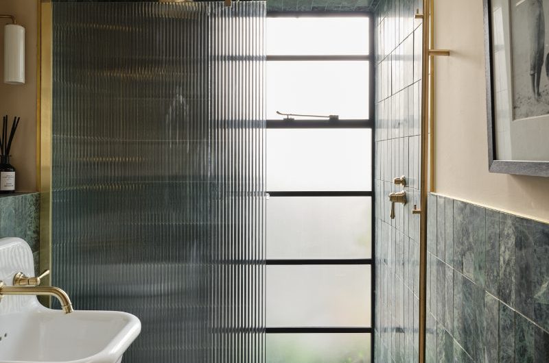 A showering space with green marble wall tiles, terrazzo flooring, a fluted panel. and tall frosted mirror.