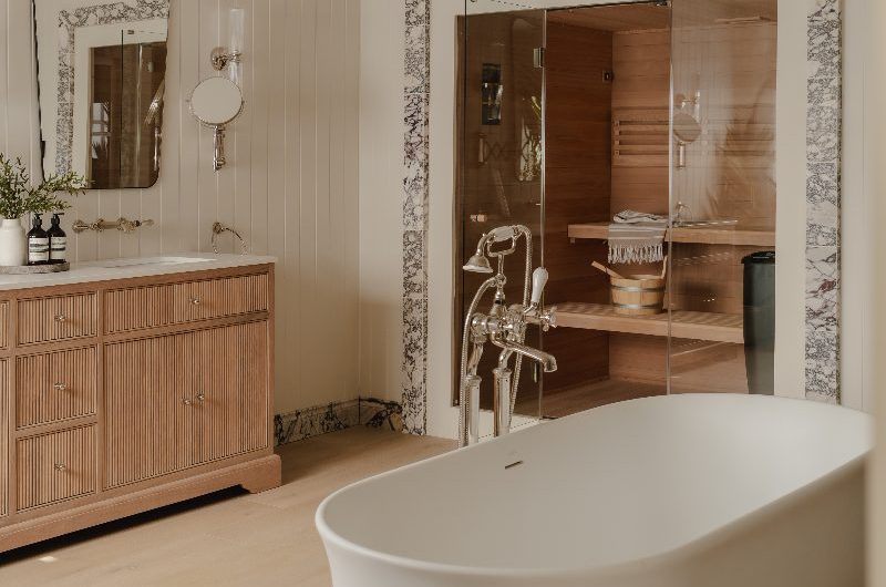 A luxury bathroom with a fluted wood vanity, a freestanding tub, and a sauna zoned by a marble trim.