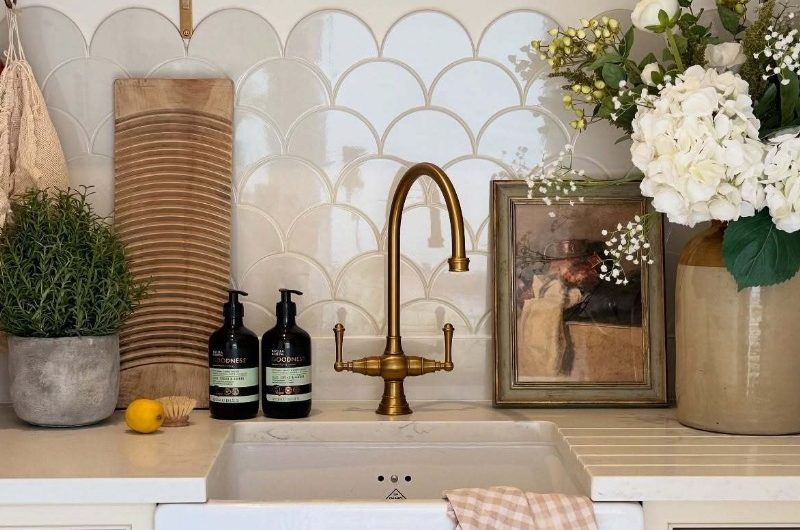Closeup at a wash zone with a butler sink, gold tap, and scalloped splashback, with a decorated open shelf above it.