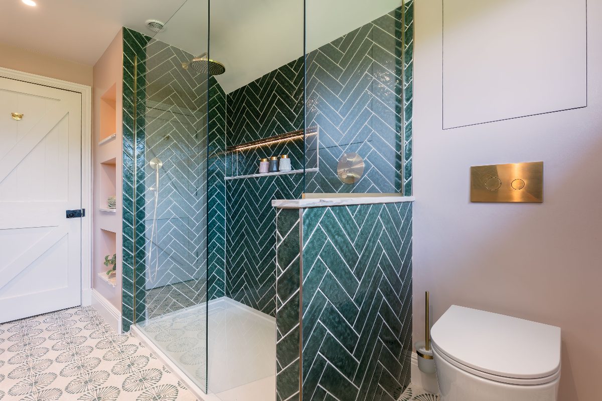 A colourful bathroom design with green herringbone tiles, pale pink paint, patterned floor tiles, white sanitaryware and sleek brassware.
