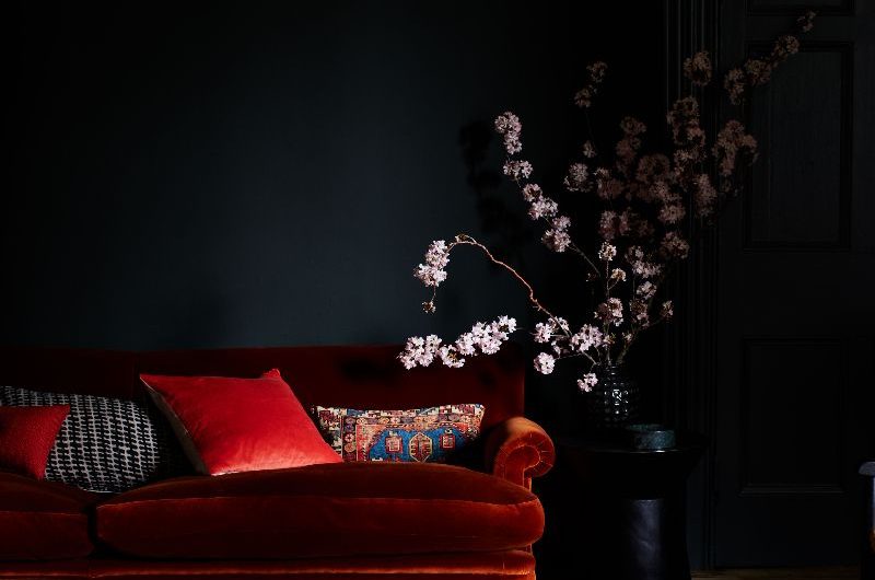 A dark living room with a dark orange velvet sofa, wood flooring, and a red patterned rug.