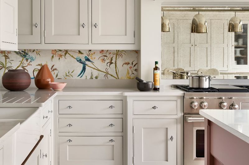 A light grey Shaker kitchen design with a stainless steel range cooker, antique mirror, botanical wallpaper as the splashback, white worktops, and large-format tiles on the floor.
