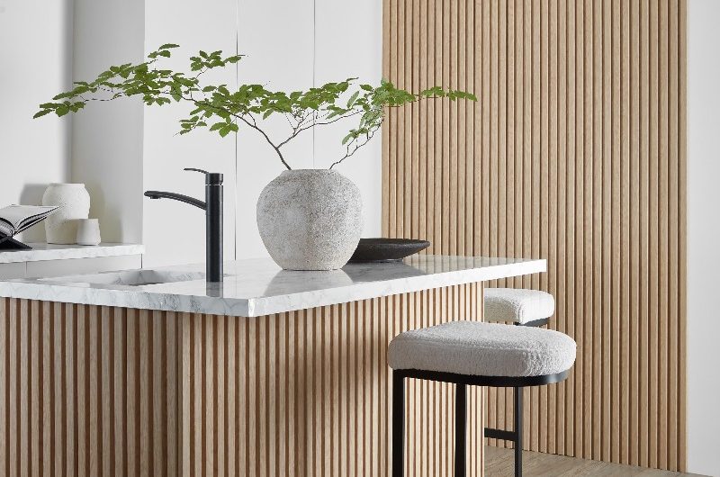 A minimalist scheme with grooved wall panelling and matching central unit with bar stools, a sink and tap, marble surface, and Japandi decor.