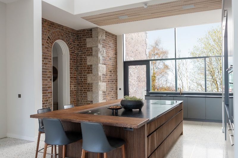 A family space with large-format floor tiles, a central island with seating, exposed brick on the walls, and tall glazing flooding the space with light.
