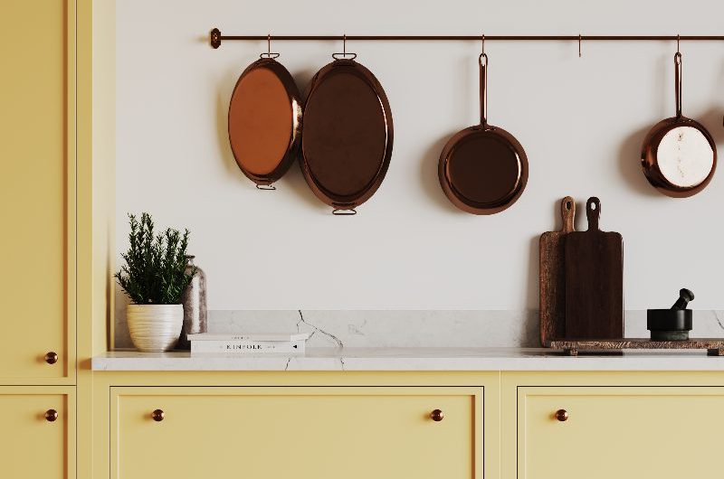 Closeup at slim Shaker cabinets painted in a warm butter yellow, with veiny work surfaces and a wall-mounted rod with hanging copper pots and pans.