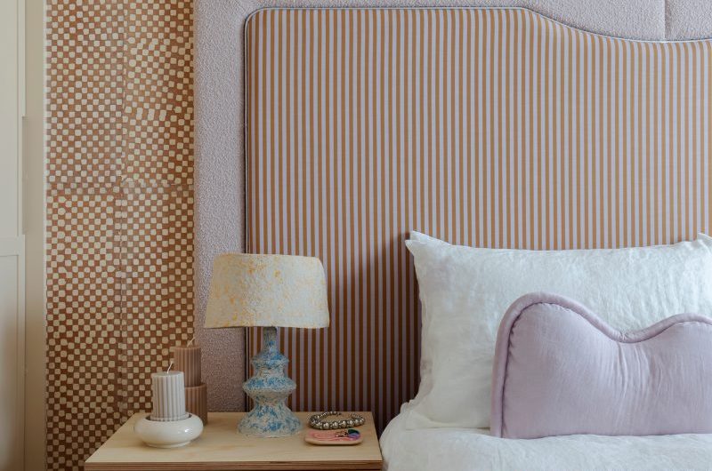 A glamorous bedroom with checkerboard wallcoverings, a striped headboard, quirky side table, and muted pastel furnishings.