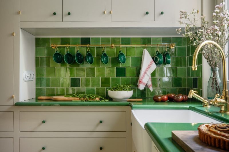 A timeless cooking space in Paris with classic cabinetry in neutral shades, green tiles, green worktops and a butler sink.