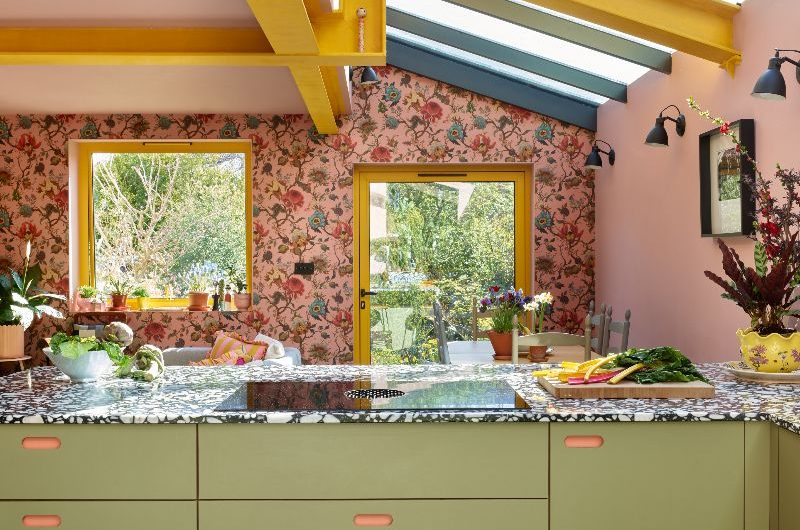 A colourful kitchen-diner with pink floral wallpaper, pink paint, yellow beams, green cabinetry and terrazzo worktops.
