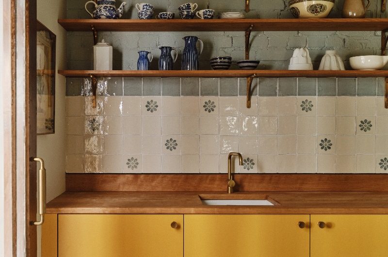 A country-chic kitchen design with yellow cabinetry, wood worktop and matching open shelving, exposed brick wall, and decorative tiles with a sage green border.