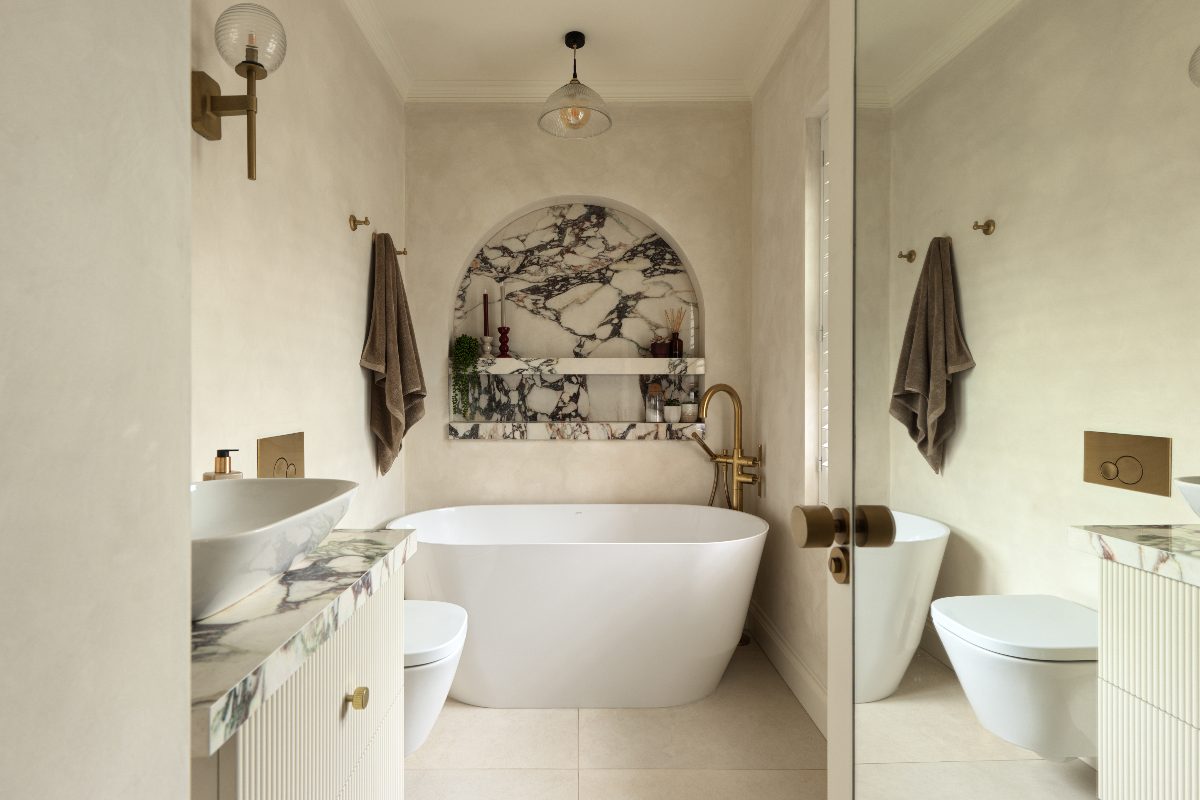 A neutral spa-like bathroom with plaster walls, a marble niche, freestanding tub, and a fluted vanity with a countertop basin.