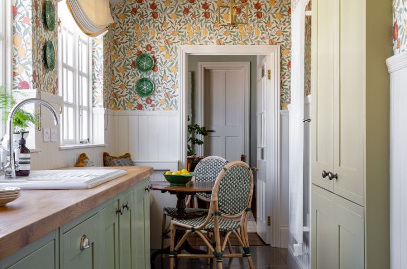 A narrow kitchen design with green Shaker units, wood worktops, a dining nook, white panelling, and citrus wallpaper on the upper half of the walls.