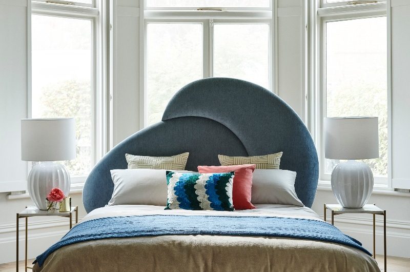 A luxurious bedroom with a quirky coloured bed frame in front of windows, colourful textiles, big table lamps, and a grand chandelier above it.