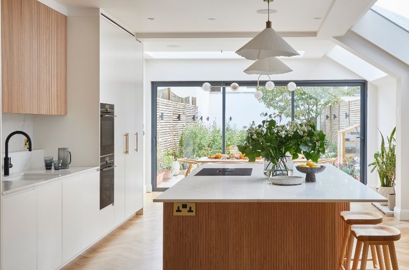 A Scandi kitchen design with a fluted island, with seating and an extractor hob, a clean and seamless run of cabinets with a fluted wood wall unit, herringbone flooring and minimalist lighting.